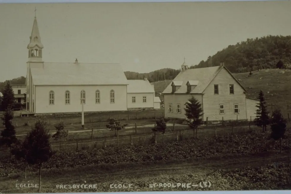 Carte postale ancienne : l’église de bois, le presbytère et l’école du village de Saint-Adolphe, les collines derrière.