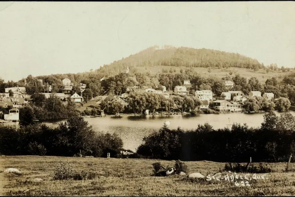 Carte postale ancienne en sépia : le village de Sainte-Adèle au bord de l’eau, une colline boisée derrière.